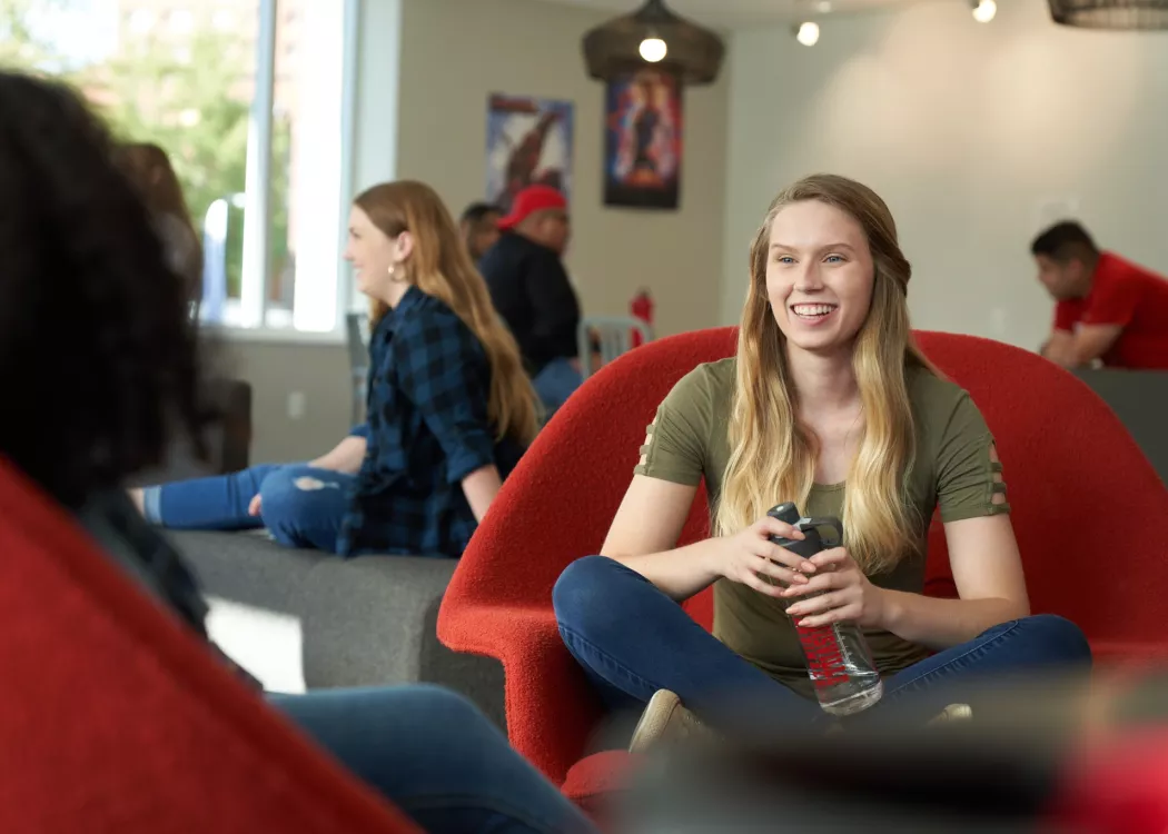 Student sitting in a red chair and talking with another student while holding a water bottle.