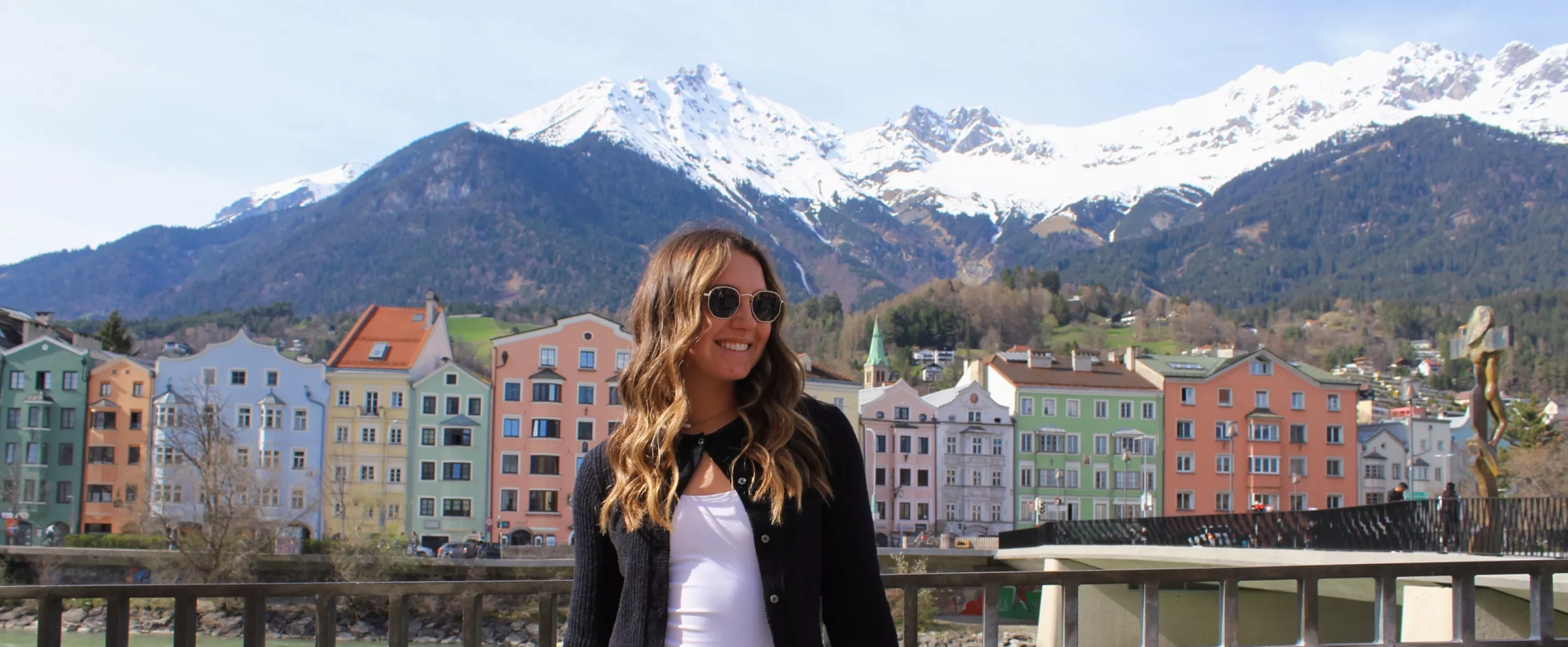 UofL student stands on bridge in front of a river, pastel colored buildings, and snow-capped mountains.